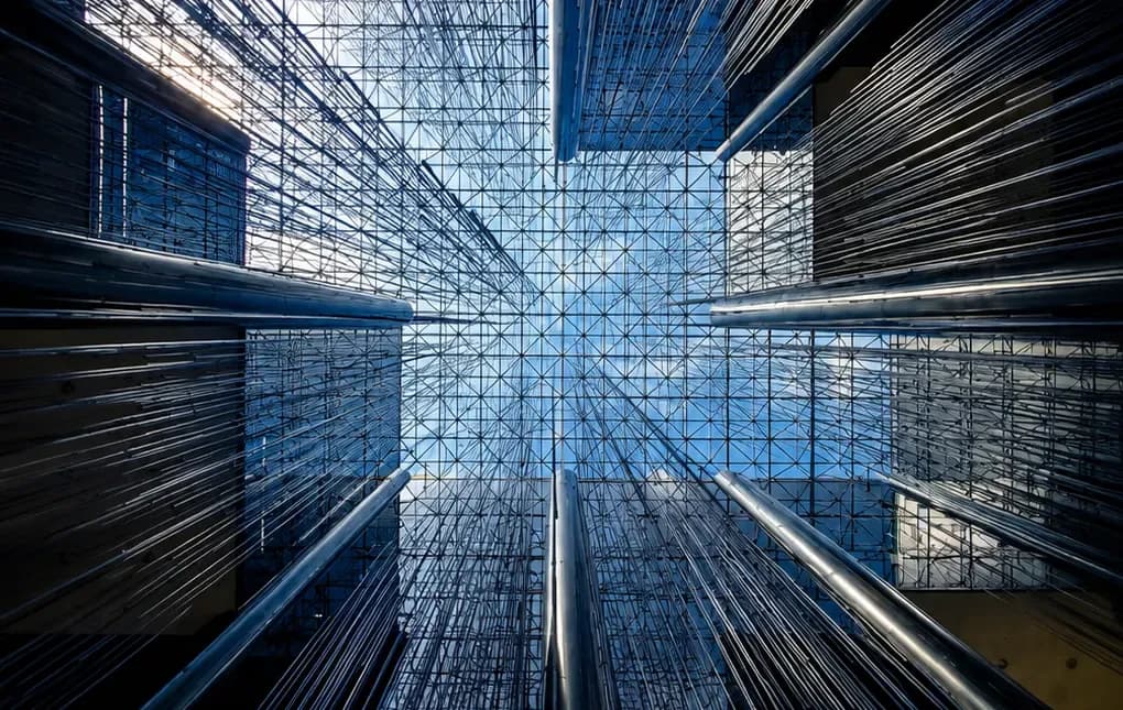Dramatic upward-looking architectural photograph inside a modern atrium, showing a geometric glass ceiling, dense metal cable or mesh structures, and reflective steel railings framed by dark building walls.