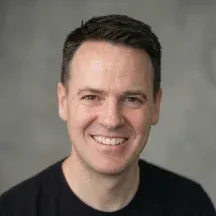 Head-and-shoulders studio portrait of a smiling adult man with short dark hair, wearing a black crew-neck shirt against a neutral gray background.